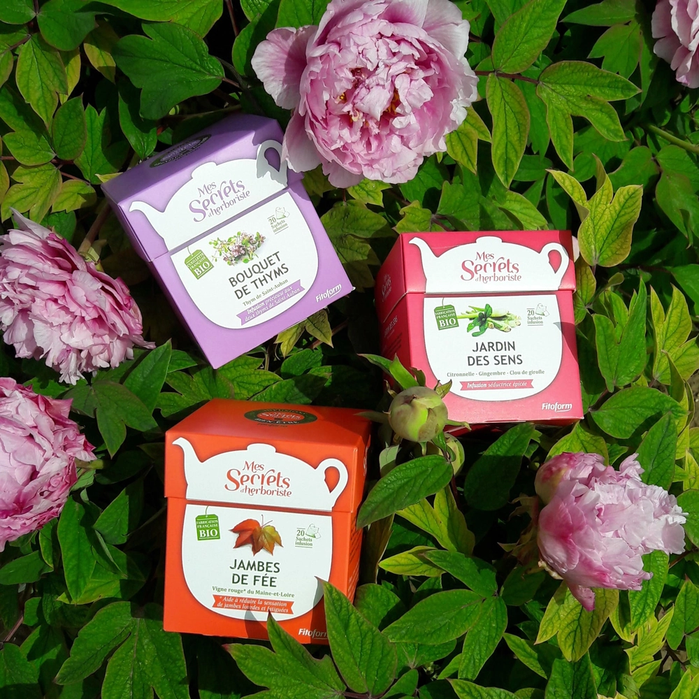 Trois boîtes d'infusions Mes Secrets d'Herboriste (Bouquet de Thyms, Jardin des Sens, Jambes de Fée) sur un parterre de pivoines roses
